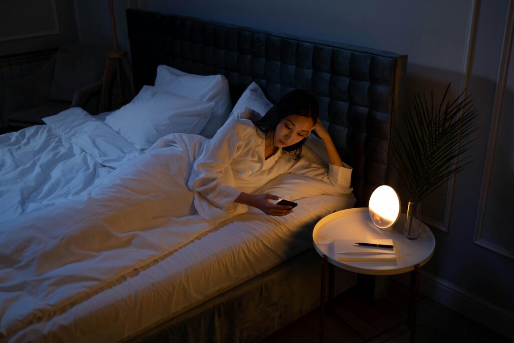An Asian woman on a bed using her phone in a dim bedroom, reflecting on insomnia or night routines.